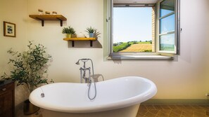 Une salle de bain avec une baignoire sur pieds, une fenêtre donnant sur un vignoble et des étagères ornées d’objets décoratifs.