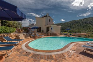A swimming pool with lounge chairs, a stone wall, and a building with a tiled roof.