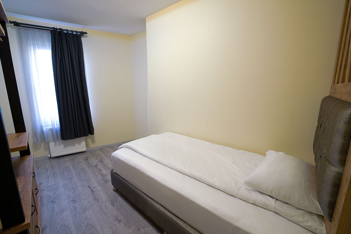 A single bed with a headboard, a wooden nightstand, a radiator, and a window with curtains.