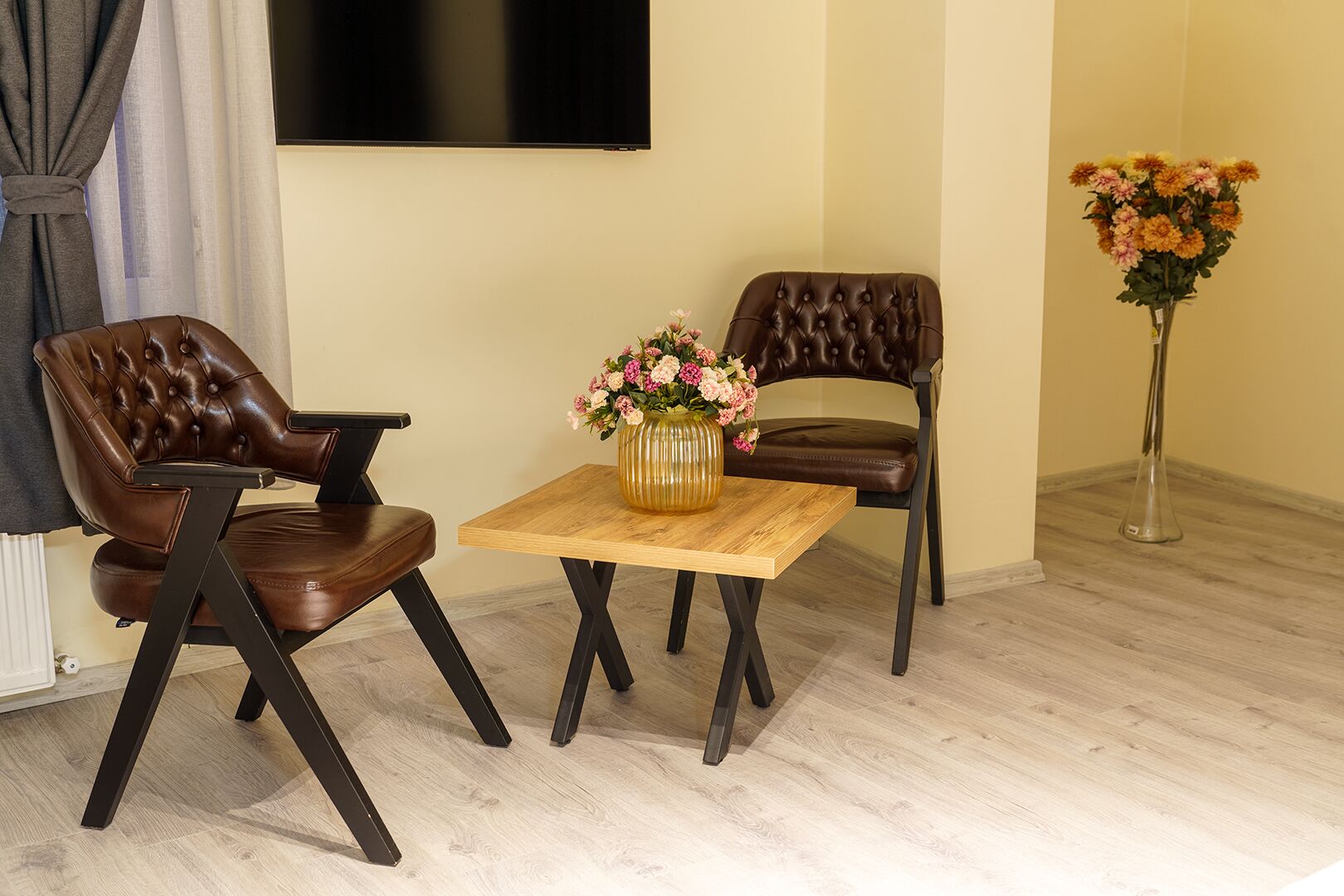 Une pièce comprenant deux fauteuils en cuir, une table en bois ornée d’un vase de fleurs, un téléviseur fixé au mur et un autre vase de fleurs posé sur le sol.