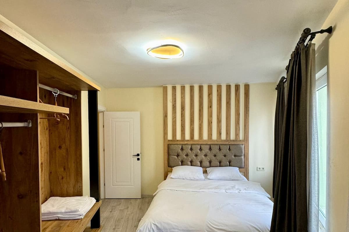A bedroom with a bed, a wooden wardrobe, a bench, a door, and a window with curtains.