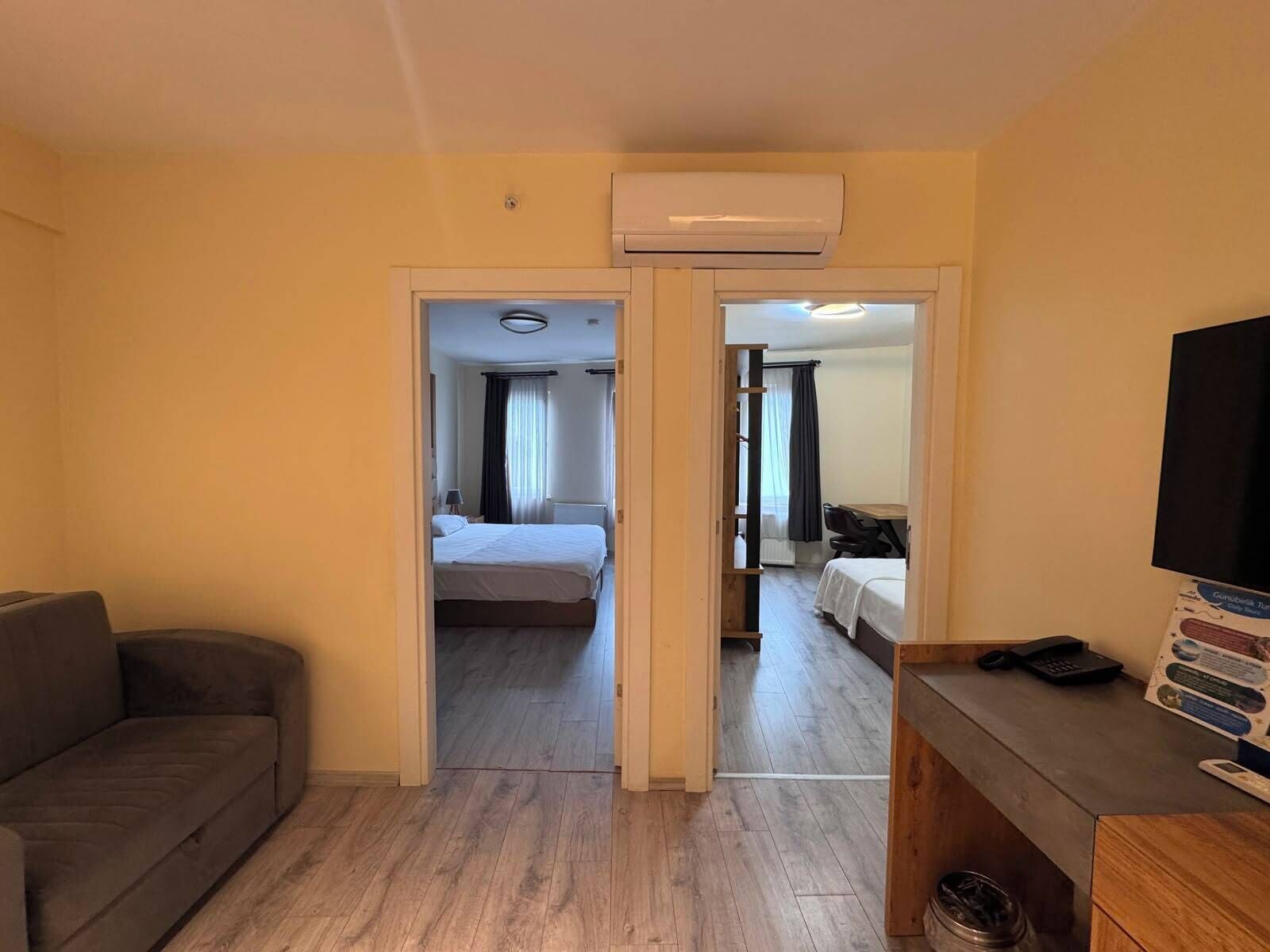 Habitación de hotel con sofá, escritorio y teléfono, y televisor. Cuenta con una cama con cabecera y cortinas, además de otra cama con disposición similar.