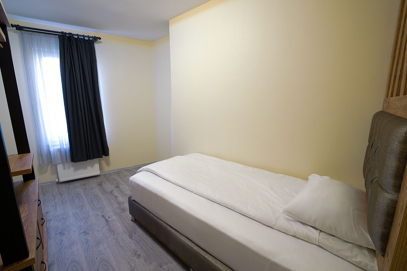 A single bed with a headboard, a wooden nightstand, a radiator, and a window with curtains.