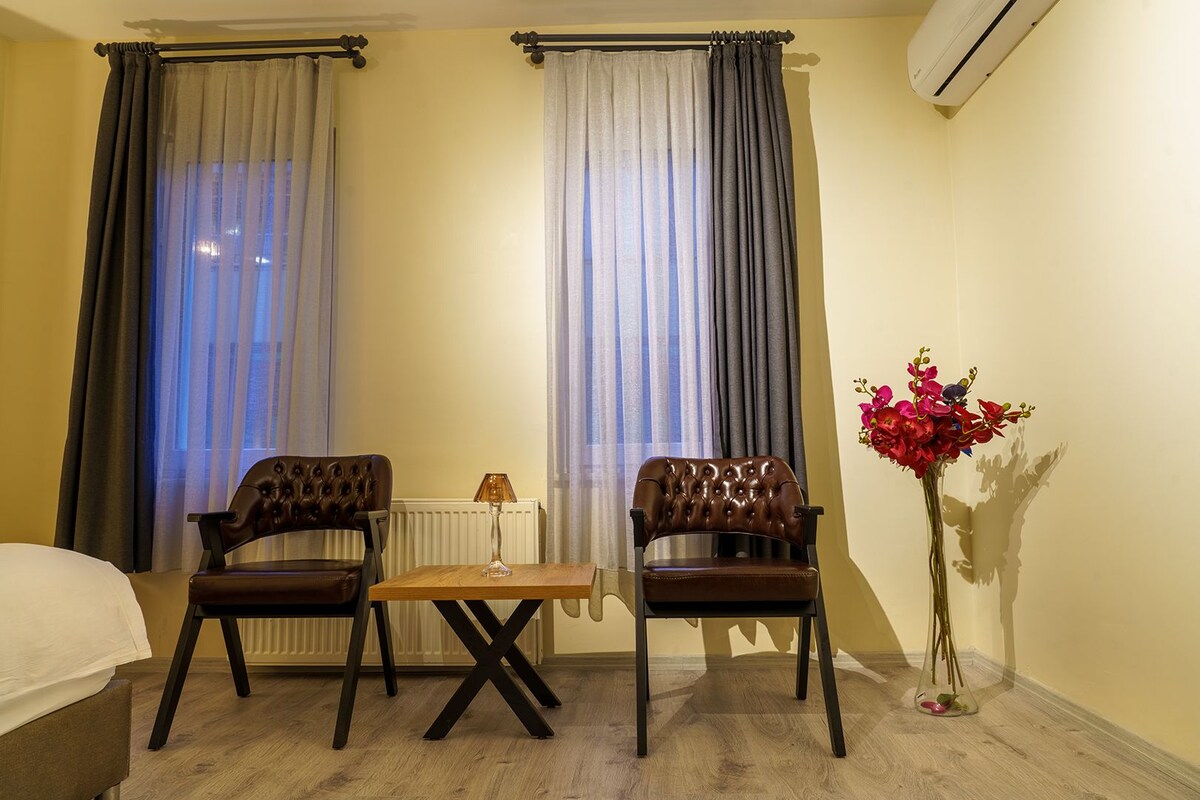 A hotel room with two leather chairs, a small wooden table, a bed, and a vase of flowers.