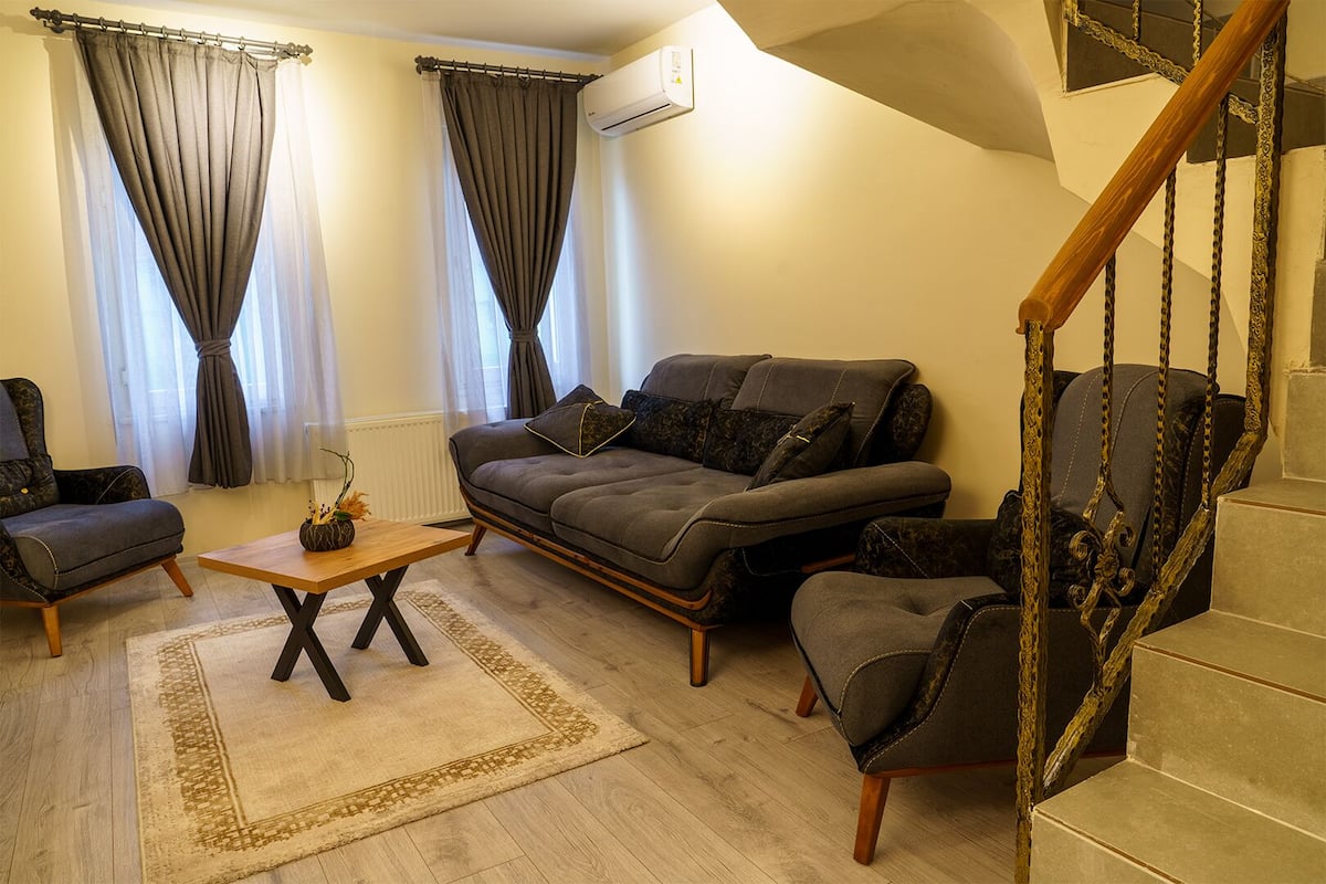 Una sala de estar con sofá, sillón y una mesita. Se observa una escalera con barandilla de madera.