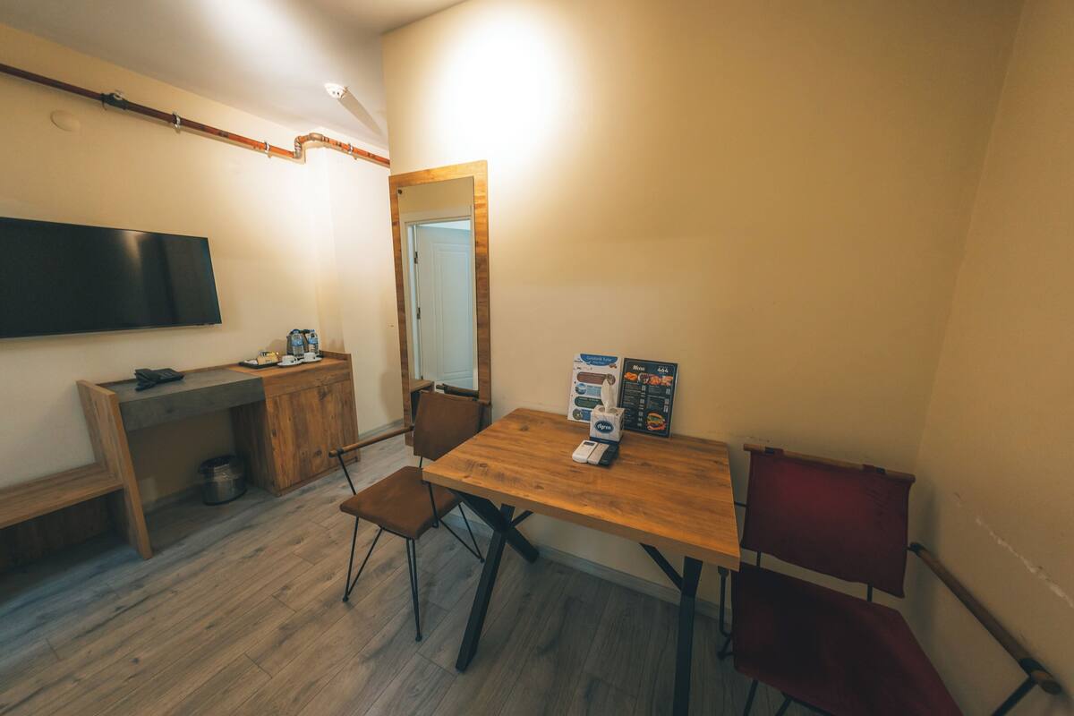 Una habitación pequeña y bien iluminada con una mesa de madera, dos sillas, un televisor montado en la pared y una puerta que da a otra habitación.