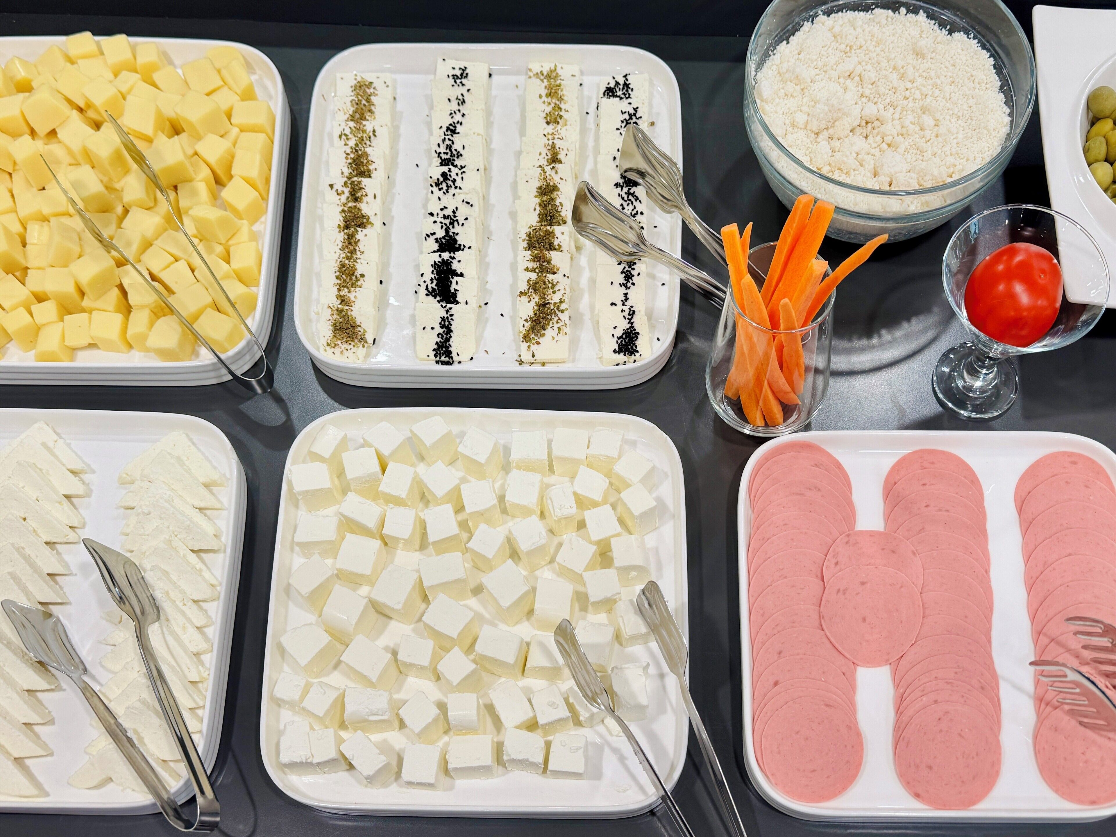 Una mesa de buffet con diversos alimentos, incluyendo queso en cubos, carnes en rodajas y verduras.