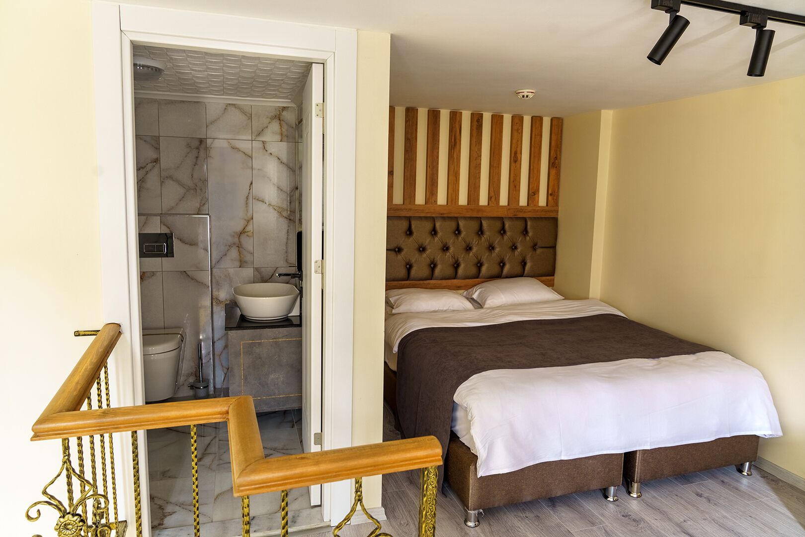 Un dormitorio con cama, cabecera de madera, baño con lavamanos y retrete de mármol, y escalera con barandilla dorada.
