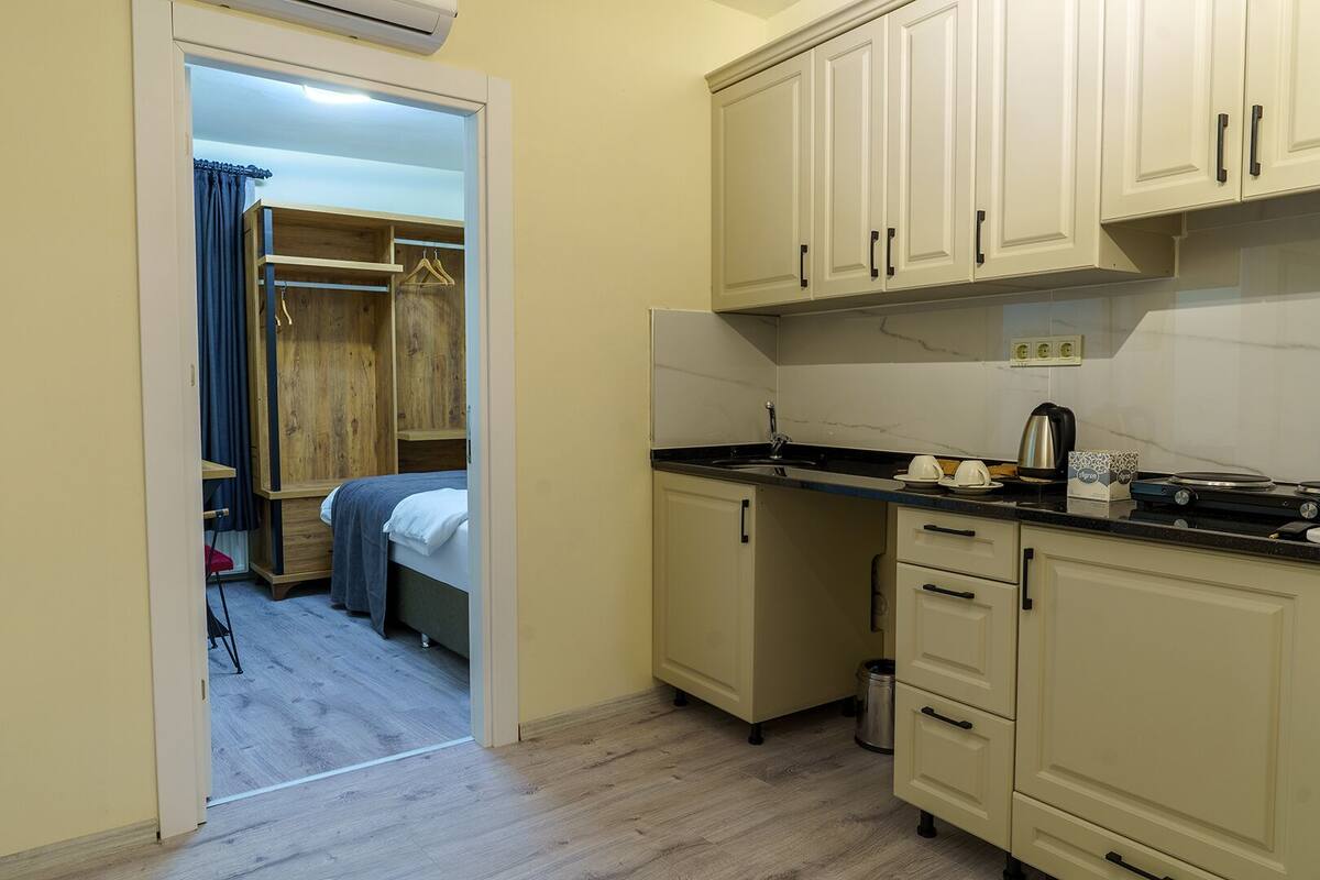 Cocina compacta con un lavabo, estufa y alacenas. Se ve una cama y una pequeña mesa en la habitación contigua.