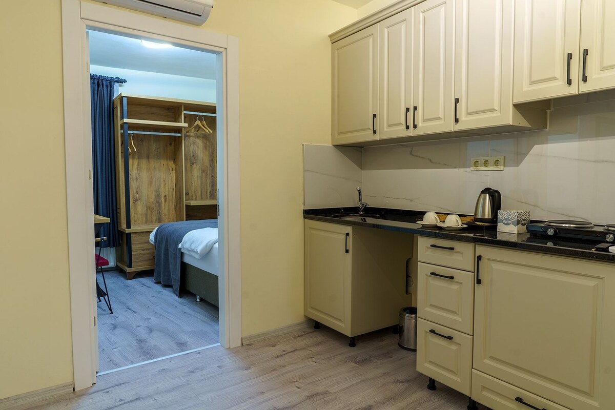 A compact kitchen with a sink, stove, and cabinets. A bed and a small table are visible in the adjacent room.