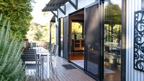 A modern outdoor dining area with a wooden deck, a table with chairs, and a glass door leading to an interior space. - Sutton Lodge (Daylesford)
