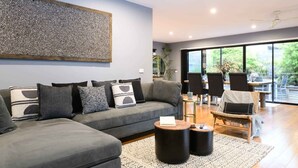 A modern living room with a grey sofa, black and white cushions, a round coffee table, and a large window offering a view of greenery. - Sutton Lodge (Daylesford)