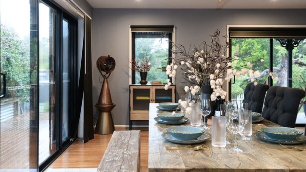 A dining area with a wooden table set for six, large windows, and a view of greenery outside. - Sutton Lodge (Daylesford)