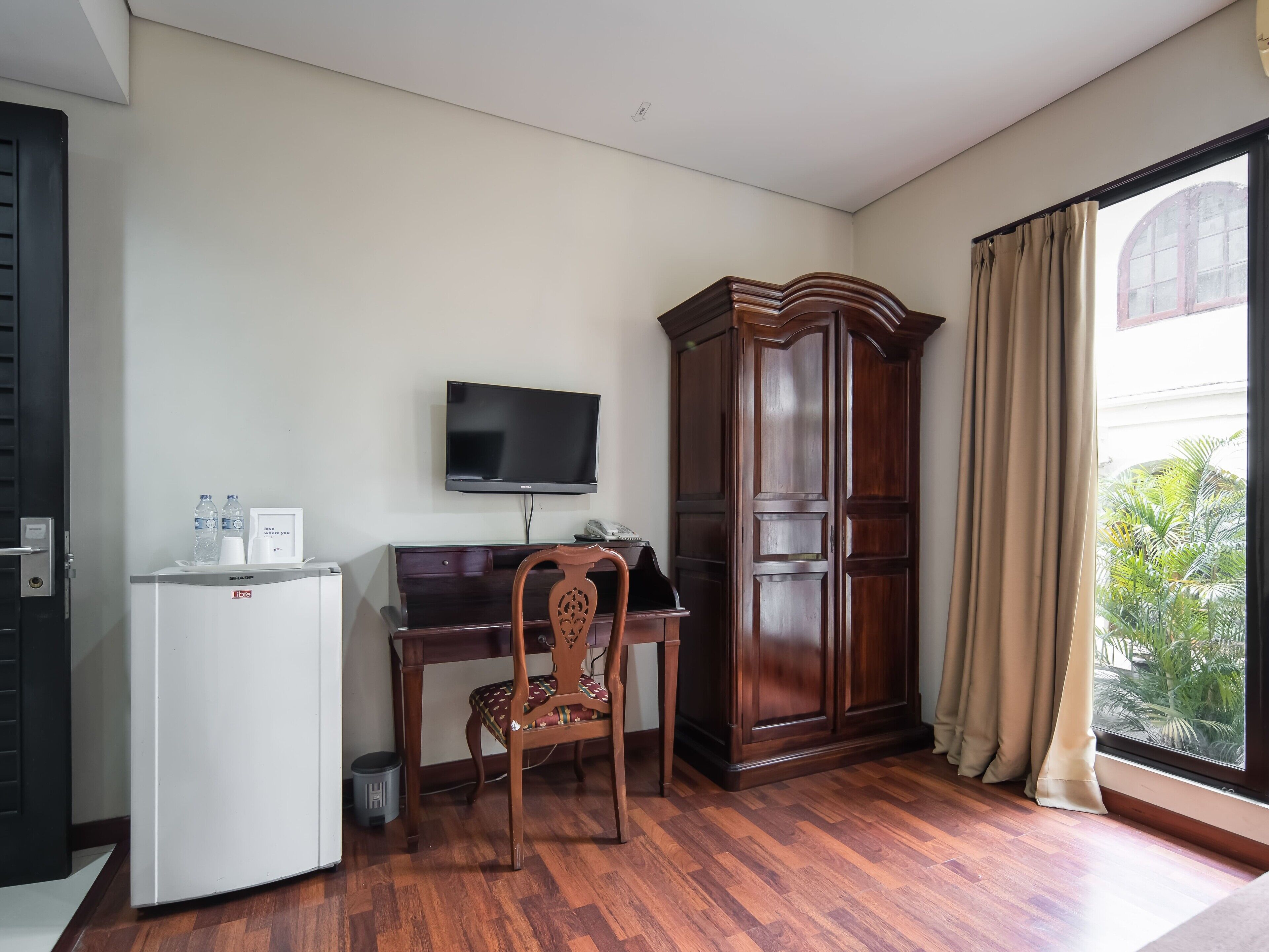 A hotel room with a wooden desk, a television mounted on the wall, a minibar, and a large window with curtains.
