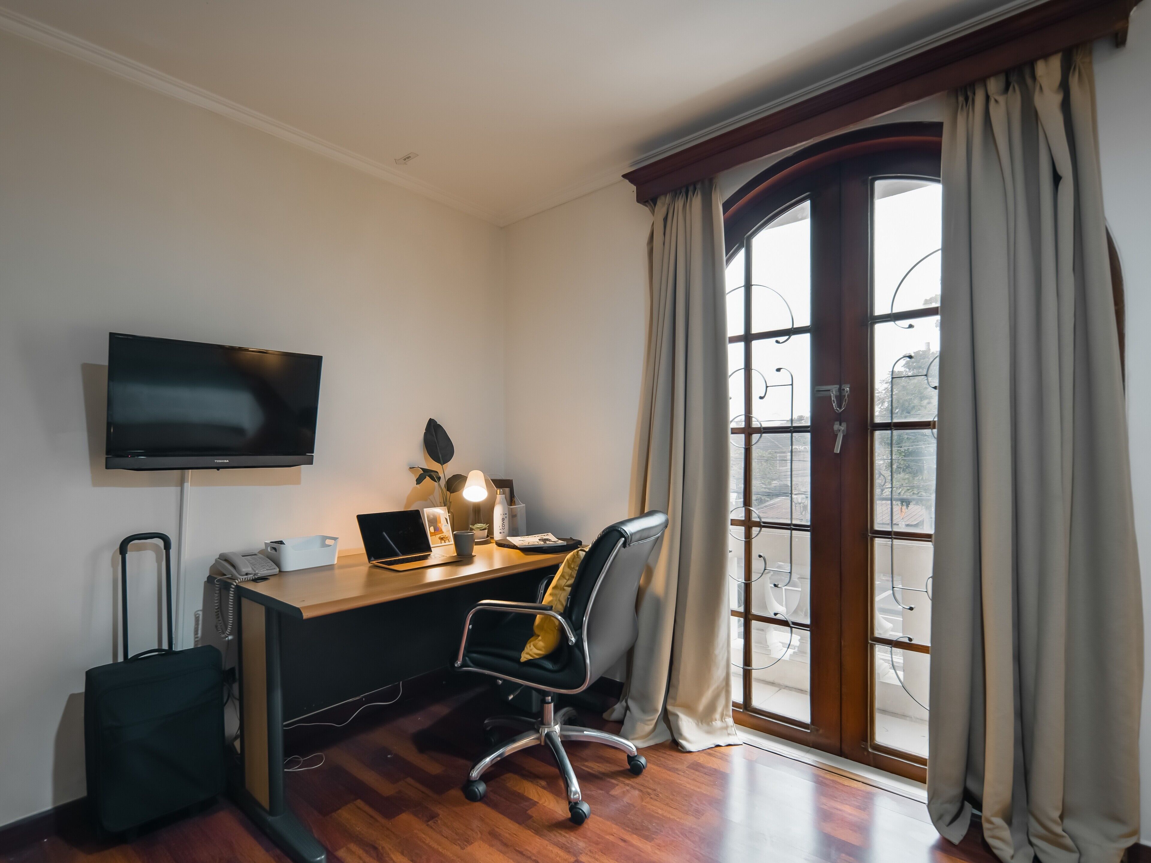 A hotel room with a wooden desk, a chair, a television, a window with curtains, and a view of the outside.