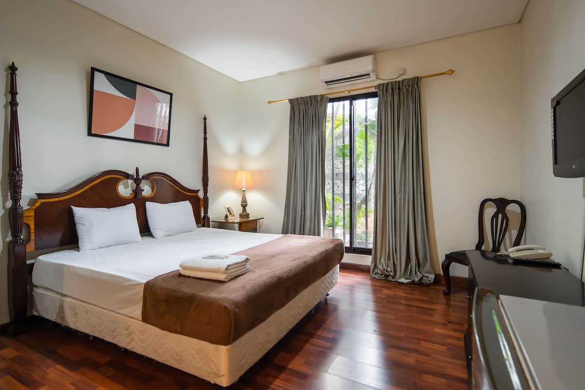 A hotel room with a large bed, a bedside table with a lamp, a desk with a phone, and a window with curtains.