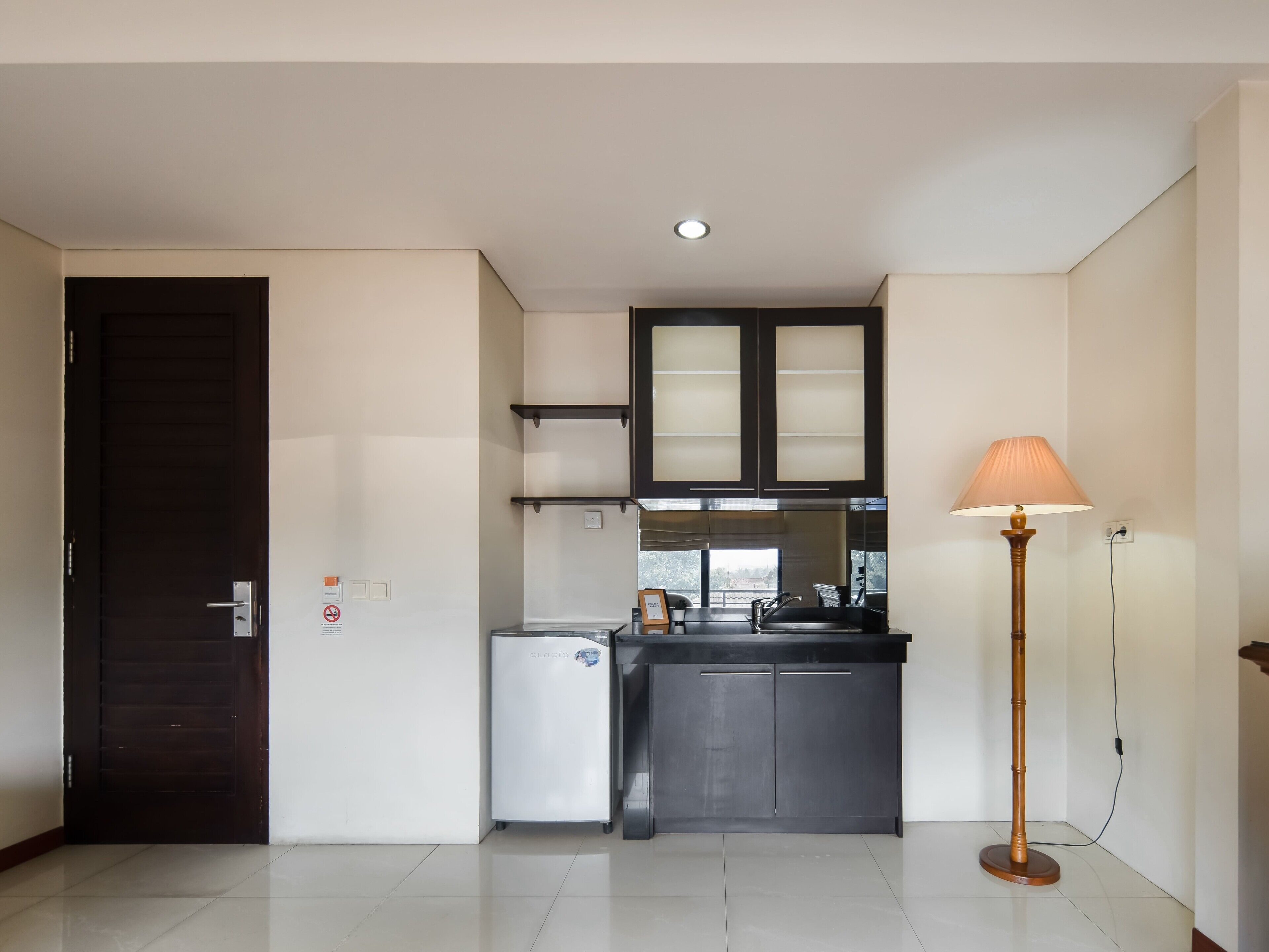 A compact kitchen with a refrigerator, a sink, and cabinets. There is a door on the left and a floor lamp on the right.