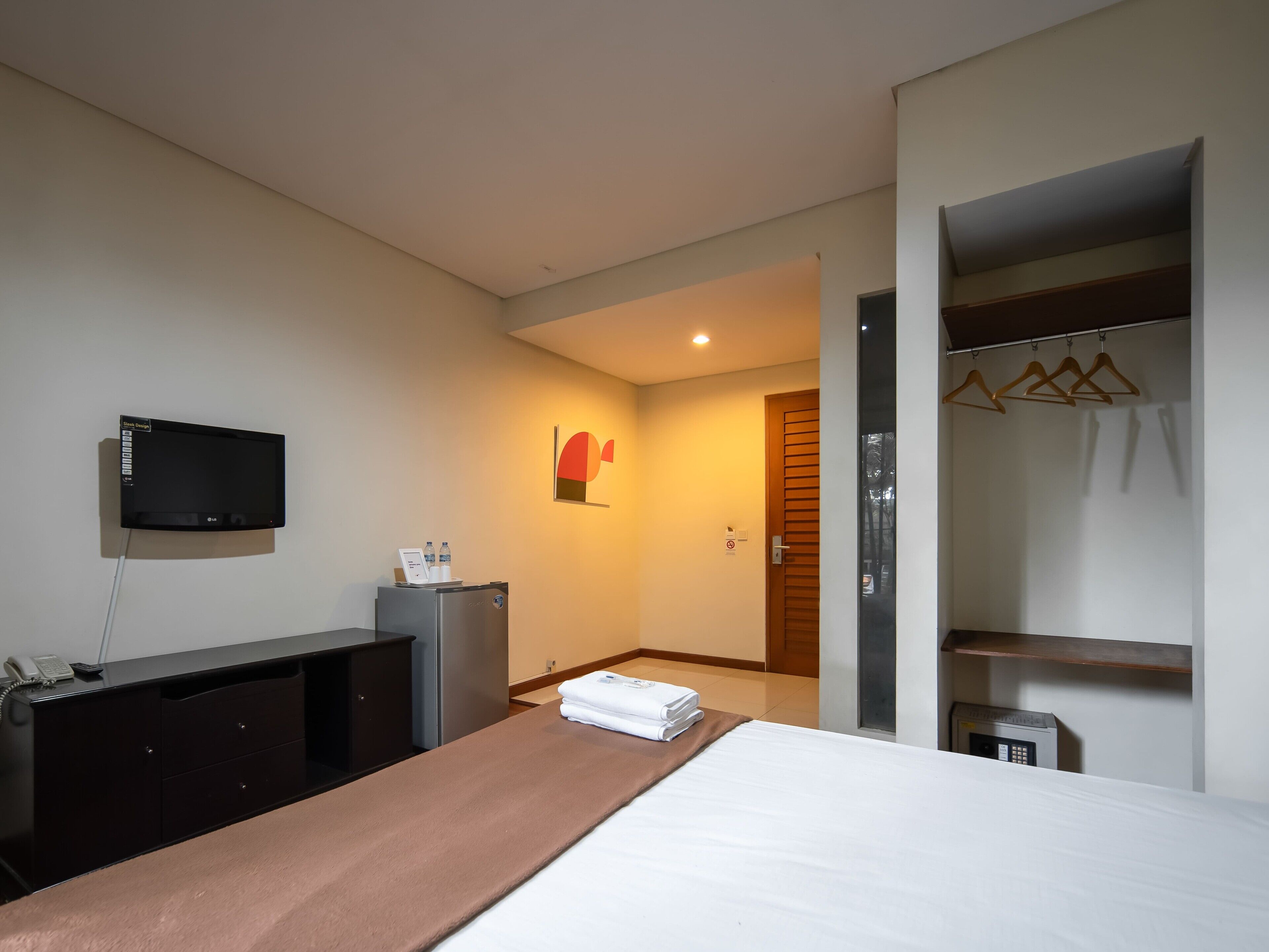 A hotel room with a bed, a desk with a computer, a TV, a coat rack, and a door leading to another room.