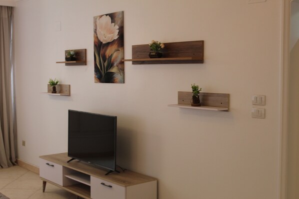 A modern living room with a flat-screen TV on a wooden TV stand, surrounded by minimalist decor including wall-mounted shelves with plants.