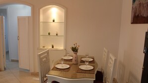 Un'area pranzo con un tavolo dal piano in vetro, sedie bianche e una piccola mensola con oggetti decorativi.