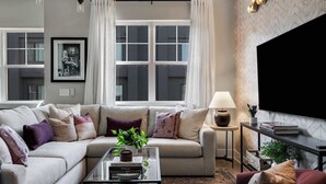 A modern living room with a sofa, coffee table, and armchair. There is a TV on the wall, a lamp, and a window with curtains. - Wildflower - Rooftop Hot Tub - Game Room (Nashville)