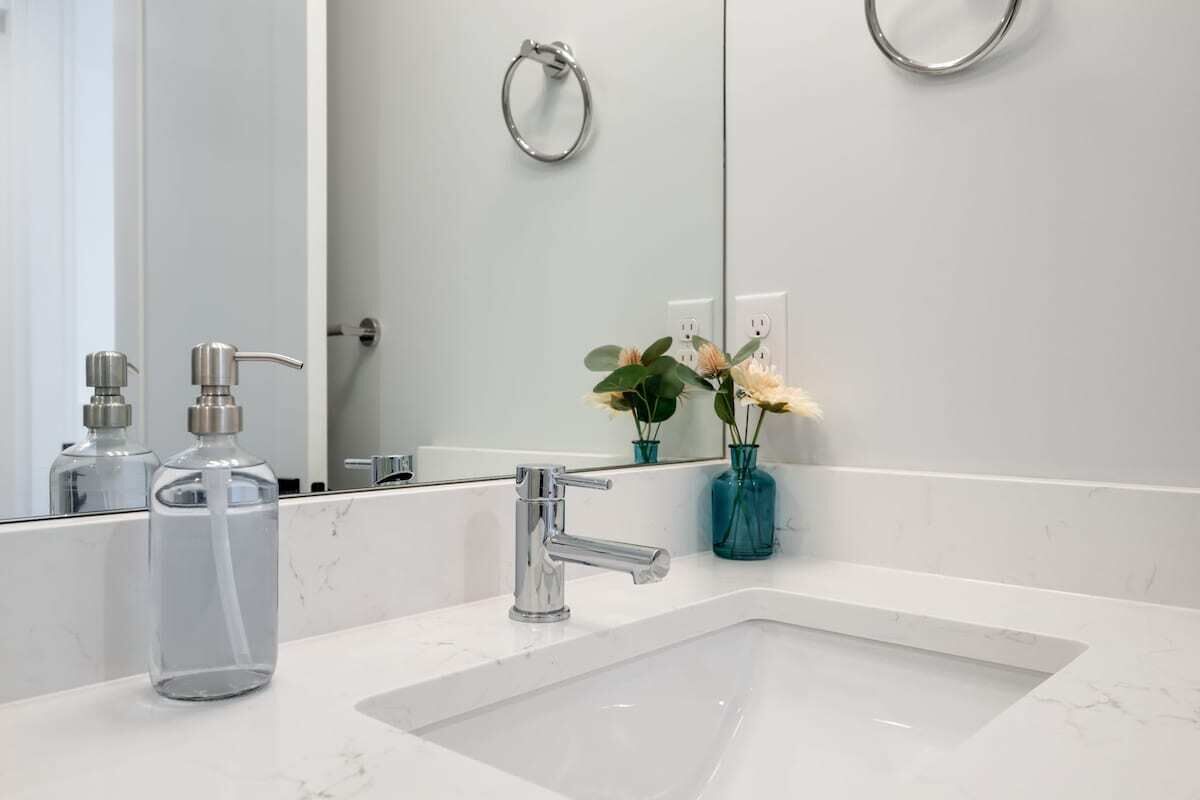 A bathroom with a white sink, a modern faucet, and a mirror reflecting the room. There are two clear glass bottles on the sink counter.