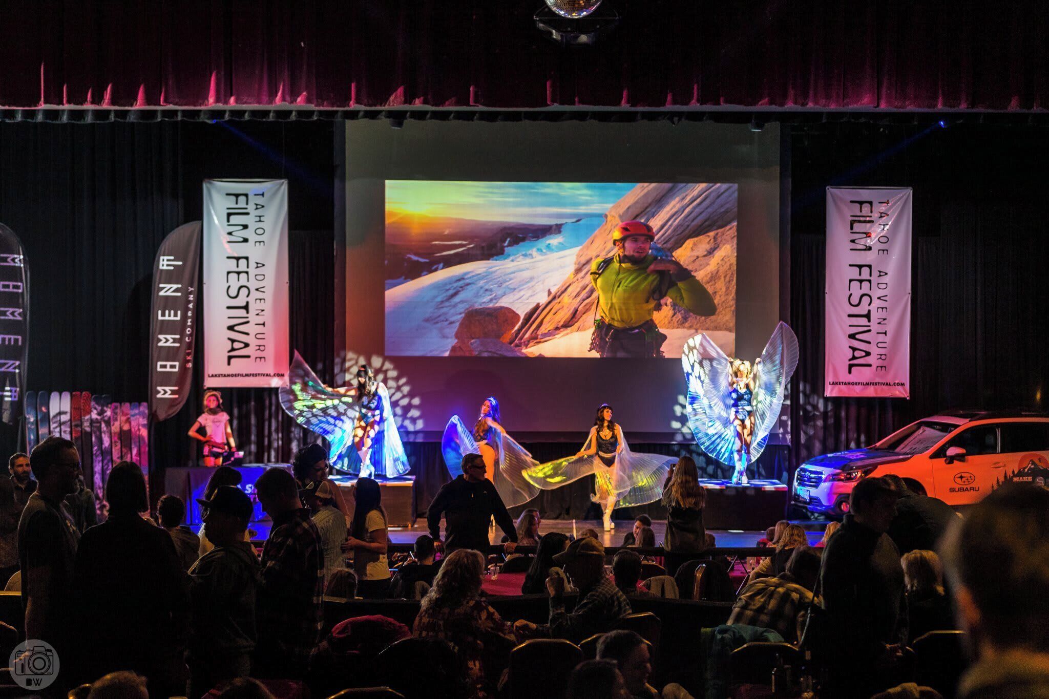 A stage with a large screen displaying a mountainous landscape, performers in costumes, and banners for the Tahoe Adventure Film Festival.
