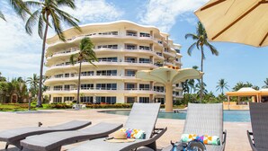 A hotel with a pool area featuring lounge chairs, a palm tree, and a large building in the background. - Gorgeous Two-Story Penthouse Private Community on the Beach! (Flamingos)