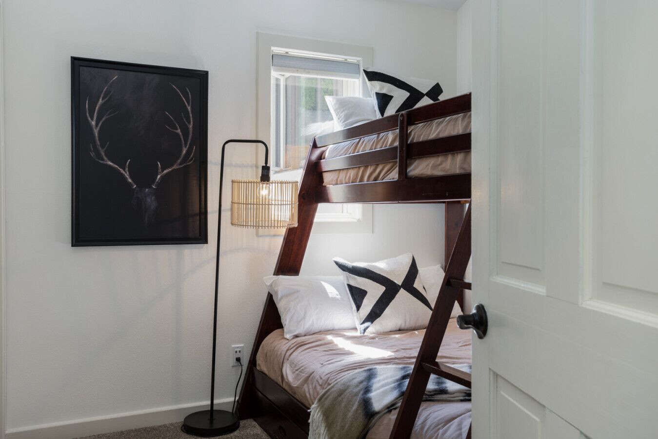 Una cama litera con cabecera, una lámpara de noche, una obra de arte con astas de venado, y una ventana con persianas.