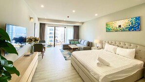 A hotel room with a large bed, a sofa, a desk, and a television. There is a balcony with a view of buildings. - Wise Stay Gold Coast Apartment (Nha Trang)