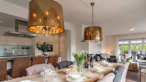 A modern dining room with a long table set for six, a kitchen in the background, and large windows offering a view of greenery outside. - Exclusive chalet with sauna and cinema seats (Mauterndorf)