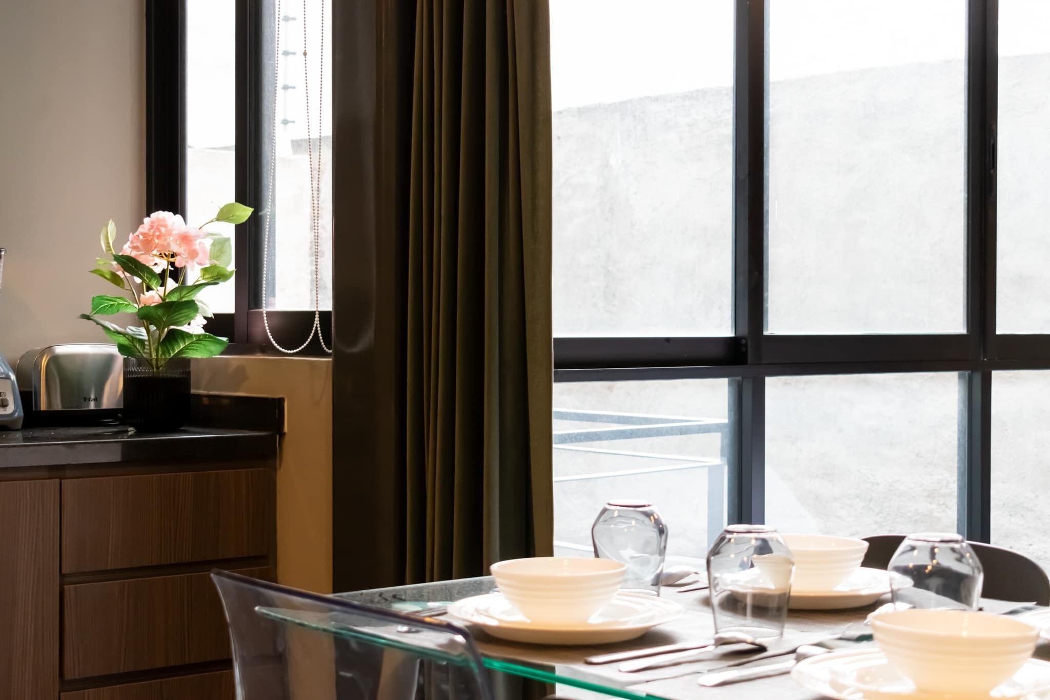 A dining area with a table set for four, a vase with flowers, and a view through large windows.