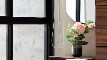 A bedroom with a large window, a bedside mirror, a wooden dresser, and a potted plant with flowers.