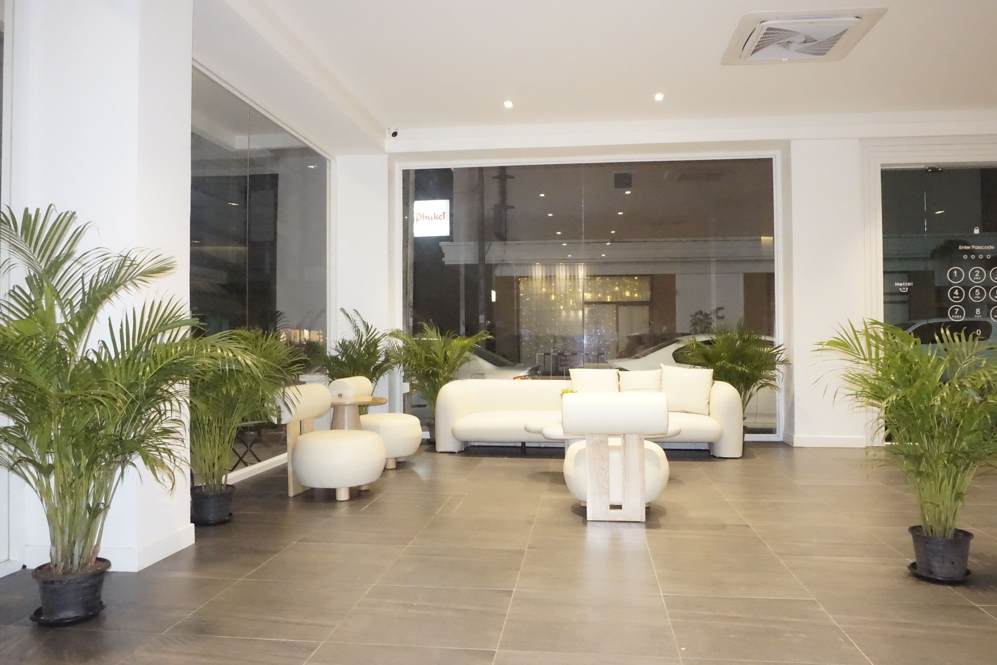 A spacious living area with a modern white sofa set, multiple potted plants, and a large window offering a view of the cityscape.