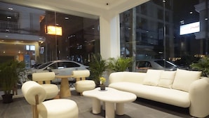 Um interior moderno com sofá, cadeiras e mesa de centro. Há plantas em vasos e vista para a rua.