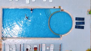 Vista aérea de uma piscina retangular com uma seção circular, cercada por cadeiras de descanso e uma grade ao redor da piscina.