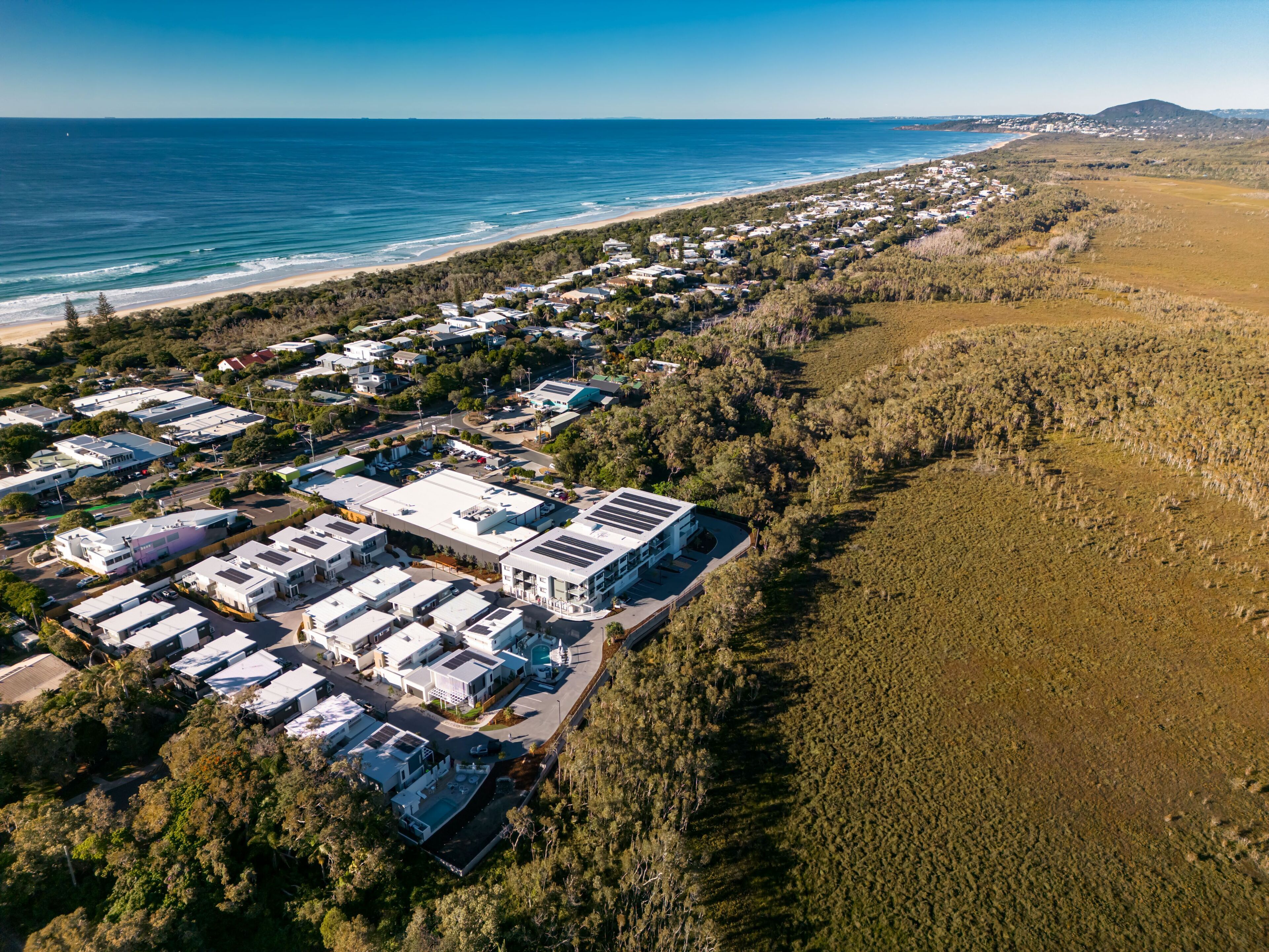 An aerial view of a coastal residential area with houses, a beach, and a hilly landscape in the background.