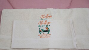 A folded white towel with embroidered text and a logo on a pink surface. - Village Boutique Hotel (Khenchara)