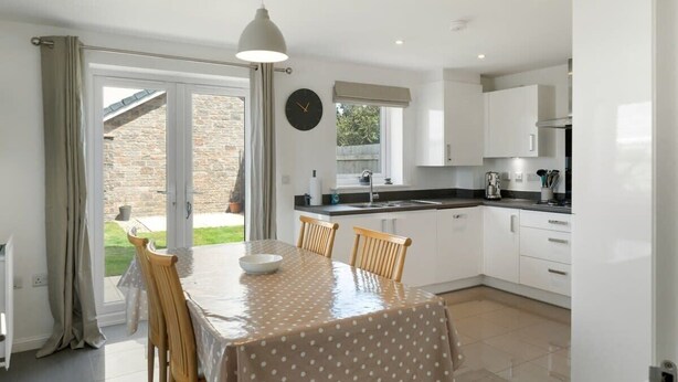 A modern kitchen with white cabinets, a dining table with chairs, and a large window. - Modern Home with Parking, nr Saunton Sands (Barnstaple)