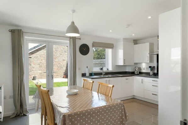 A modern kitchen with white cabinets, a dining table with chairs, and a large window. - Modern Home with Parking, nr Saunton Sands (Barnstaple)
