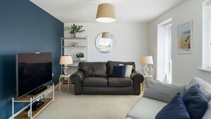 A modern living room with a dark blue accent wall, a grey sofa, a brown leather couch, a flat-screen TV on a wooden stand, and a round mirror on the wall. - Modern Home with Parking, nr Saunton Sands (Barnstaple)