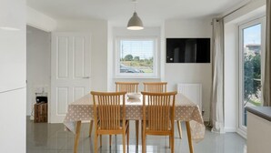 A dining area with a table and chairs, a window with blinds, a television mounted on the wall, and a view of the outdoors. - Modern Home with Parking, nr Saunton Sands (Barnstaple)