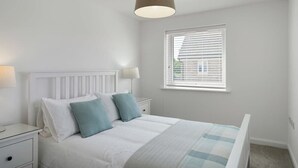 A bedroom with a white bed, two nightstands, a window with blinds, and a hanging light fixture. - Modern Home with Parking, nr Saunton Sands (Barnstaple)