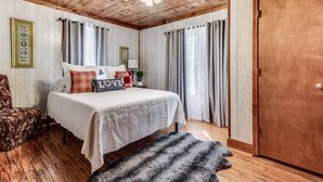 A bedroom with a wooden ceiling, a bed with a white bedspread and pillows, a wooden door, and a window with curtains.