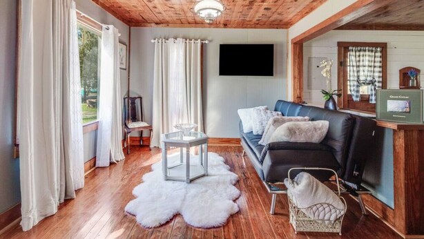 A cozy living room with a wooden ceiling, a black sofa, a glass coffee table, and a fluffy white rug.