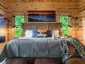 Une chambre à coucher dans une cabane en bois, avec un lit, des oreillers, une table de chevet, une lampe et un tableau représentant un coucher de soleil.