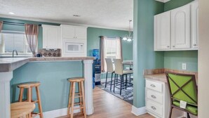 A kitchen with white cabinets, a wooden island with stools, a microwave, and a dining area with chairs. - Family Friendly Waterfront Home | Private backyard (New Orleans)