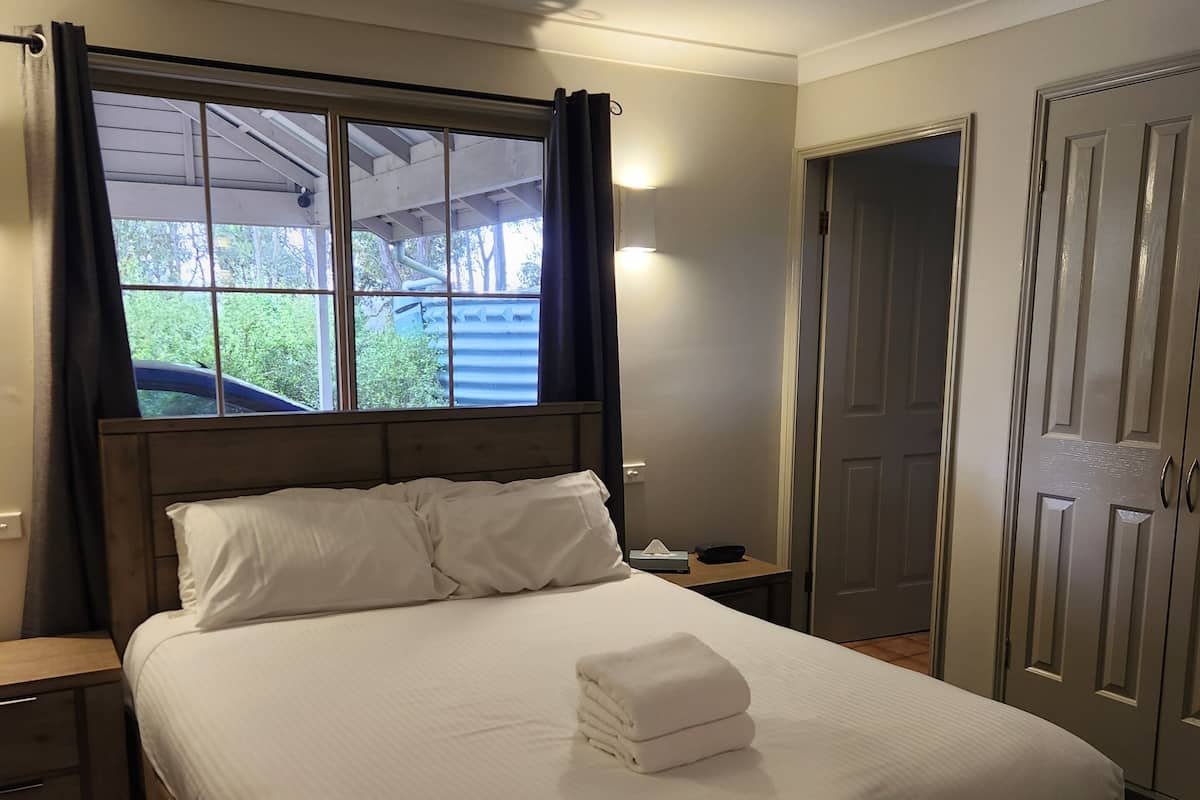 A neatly made bed with a folded towel, a bedside table with a lamp, and a window with curtains.