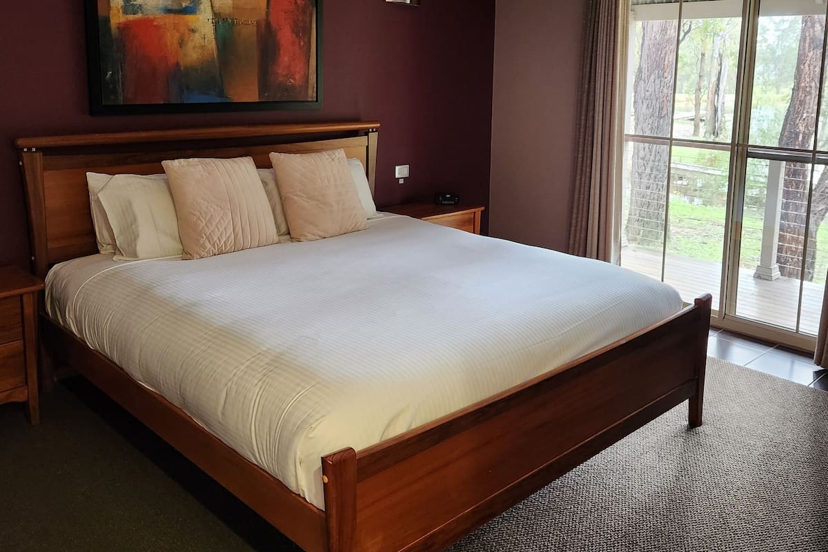 A large bed with a wooden frame and white bedding, a ceiling light, a painting on the wall, and a window with curtains.