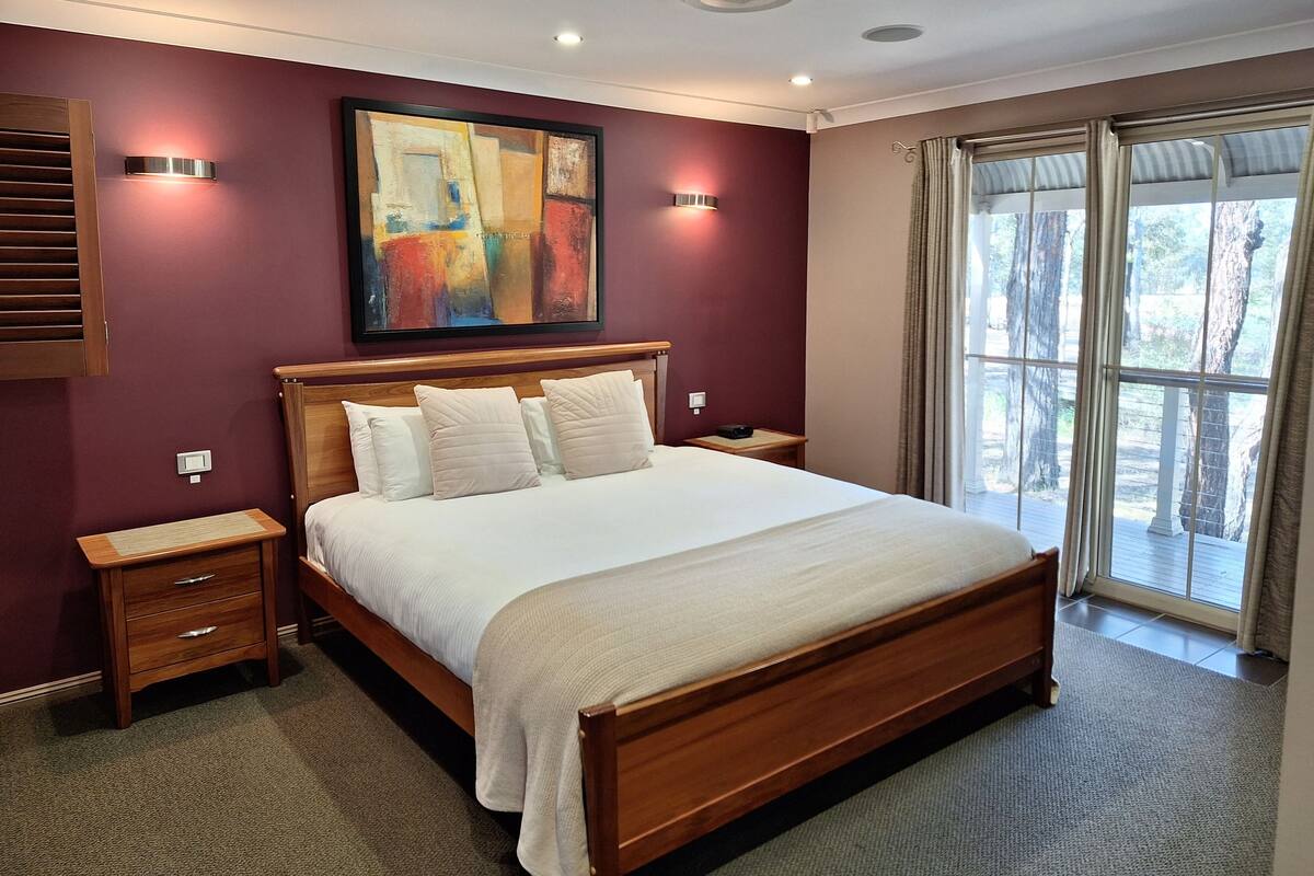 A bedroom with a large bed, wooden headboard, bedside tables, a painting on the wall, and a view of trees through the window.